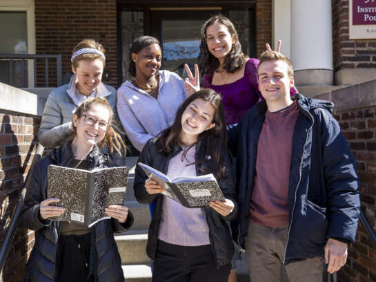 Six students outside IOP, smiling. Two students are holding composition notebooks.