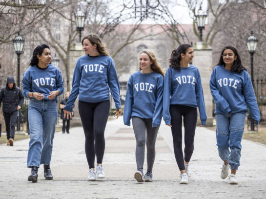 Five students walk through campus wearing blue shirts that say "Vote."